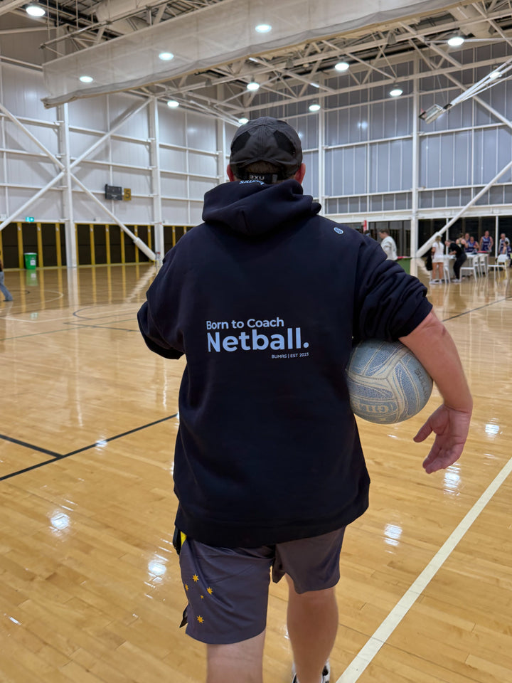 Person holding a netball in an indoor sports facility wearing a 'Born to Coach Netball' hoodie.